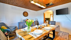 Un coin repas avec une table en bois dressée pour quatre personnes, une horloge au mur et un téléviseur à écran plat fixé au-dessus de la table.