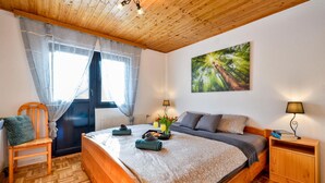 Une chambre avec un plafond en bois, un lit recouvert d’une couvre-lit à motifs floraux, une fenêtre ornée de rideaux légers, un tableau représentant une forêt et une table de chevet avec une lampe.