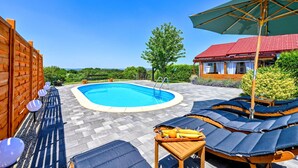 Une piscine entourée d’une clôture en bois, avec des chaises longues et des parasols, et une maison avec un toit rouge.