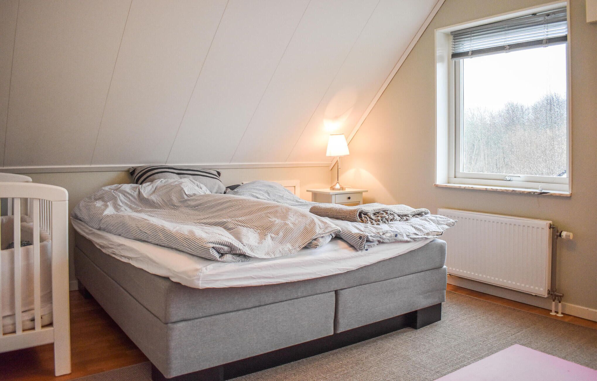 A bedroom with a grey bed, a white crib, a nightstand with a lamp, a window with a view of trees, and a radiator.