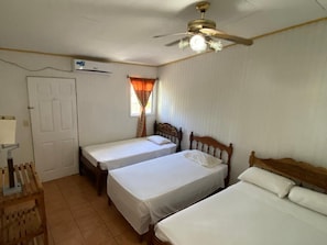 A room with two single beds, a ceiling fan, an air conditioning unit, a window with curtains, and a wooden table. - Villas Yulli (Tela)