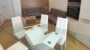Een moderne woonkamer met een glazen eettafel, twee banken, een keuken met inbouwapparatuur en een wand bekleed met hout.