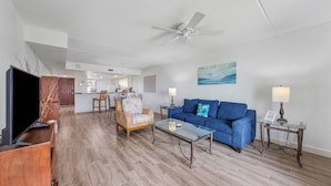 A living room with a blue sofa, a glass coffee table, a TV, and a ceiling fan. - Beach FRONT Hideaway for your Fun in the Sun!️ (South Padre Island)