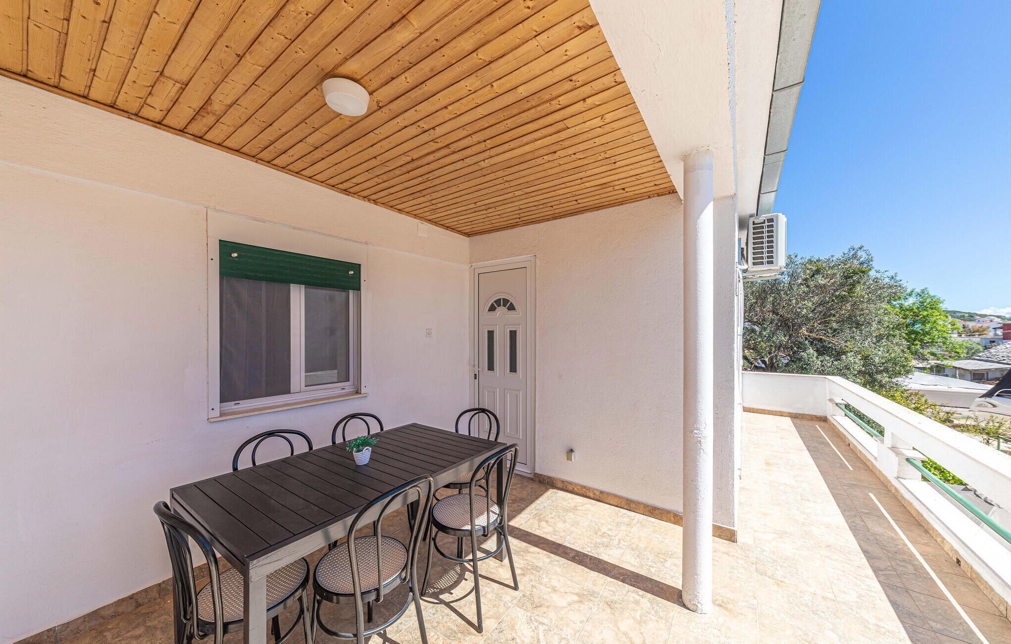 Eine überdachte Terrasse im Freien mit Holzboden, einem Tisch mit Stühlen, einem Fenster mit grüner Rollo und einer Klimaanlage.