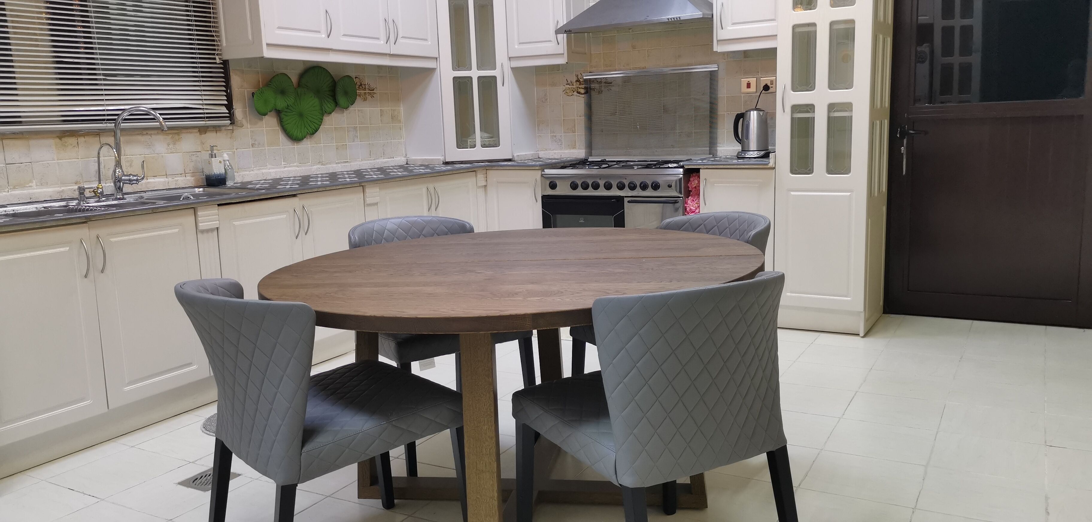 Una cocina con una mesa redonda de madera, cuatro sillas grises, armarios blancos, una estufa y un lavamanos.