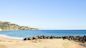 A beach with a rocky shoreline, clear blue water, and a hilly landscape in the background. - Nice apartment in Giardini Naxos with WiFi (Giardini Naxos)