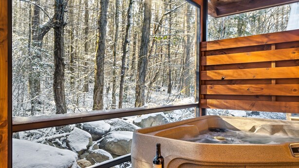 Ein Whirlpool mit einer Weinflasche und Trauben, ein Fenster mit Blick auf einen verschneiten Wald und Holzbalken.