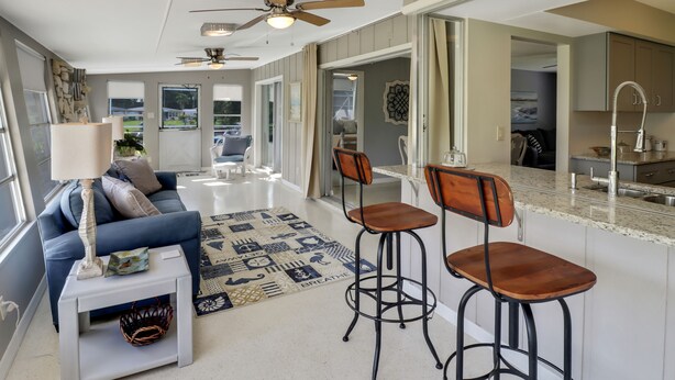 A modern living room with a blue sofa, a coffee table, and two bar stools. There is a ceiling fan, a floor lamp, and a large window providing natural light. - Canal Home to Gulf Completely Renovated, Large Pool, come to Serenity Retreat!!! (Englewood)
