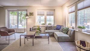 A modern living room with a sofa, armchair, and coffee table. There are two windows with blinds, a ceiling fan, and a view of the outdoors.