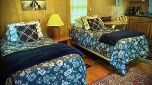 A room with two beds, a small table, a lamp, a window with blinds, and a kitchen area.