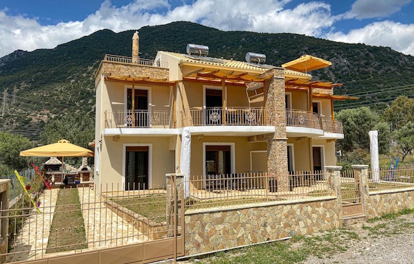 Edifici de dues plantes amb balcons, façana de pedra i un paisatge de muntanyes al fons.
