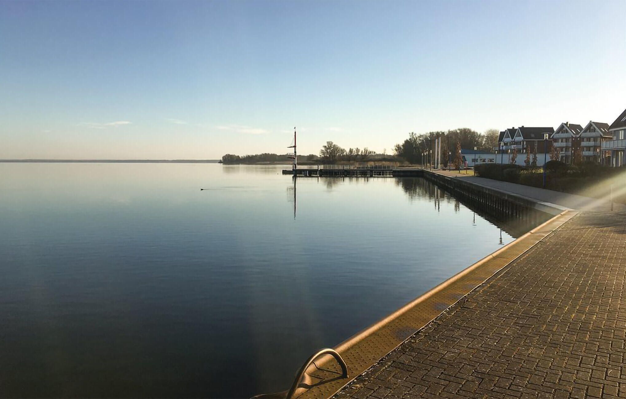 Una orilla tranquila con un paseo marítimo empedrado, un muelle y edificios residenciales al fondo.