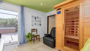Una habitación con sauna de madera, un sillón de cuero negro, una silla de madera, una planta en maceta y una pared con cuadros.