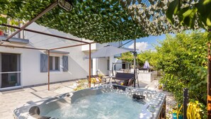 Un jacuzzi con una pérgola encima, un patio con mobiliario exterior y un jardín con diversas plantas.