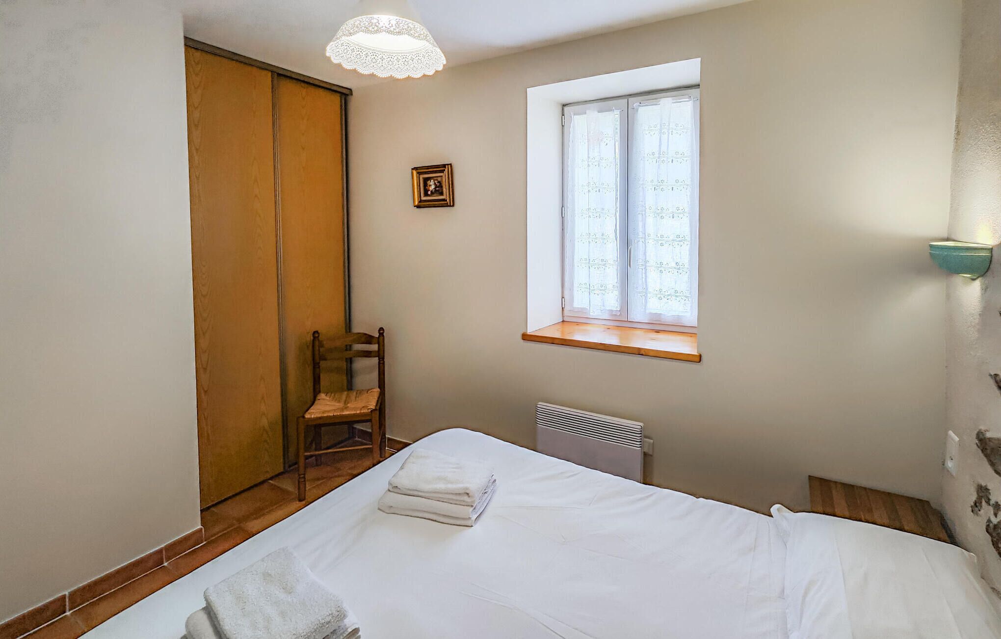Una camera da letto con un letto, una finestra con tende di pizzo, un termosifone, una sedia di legno e un armadio.
