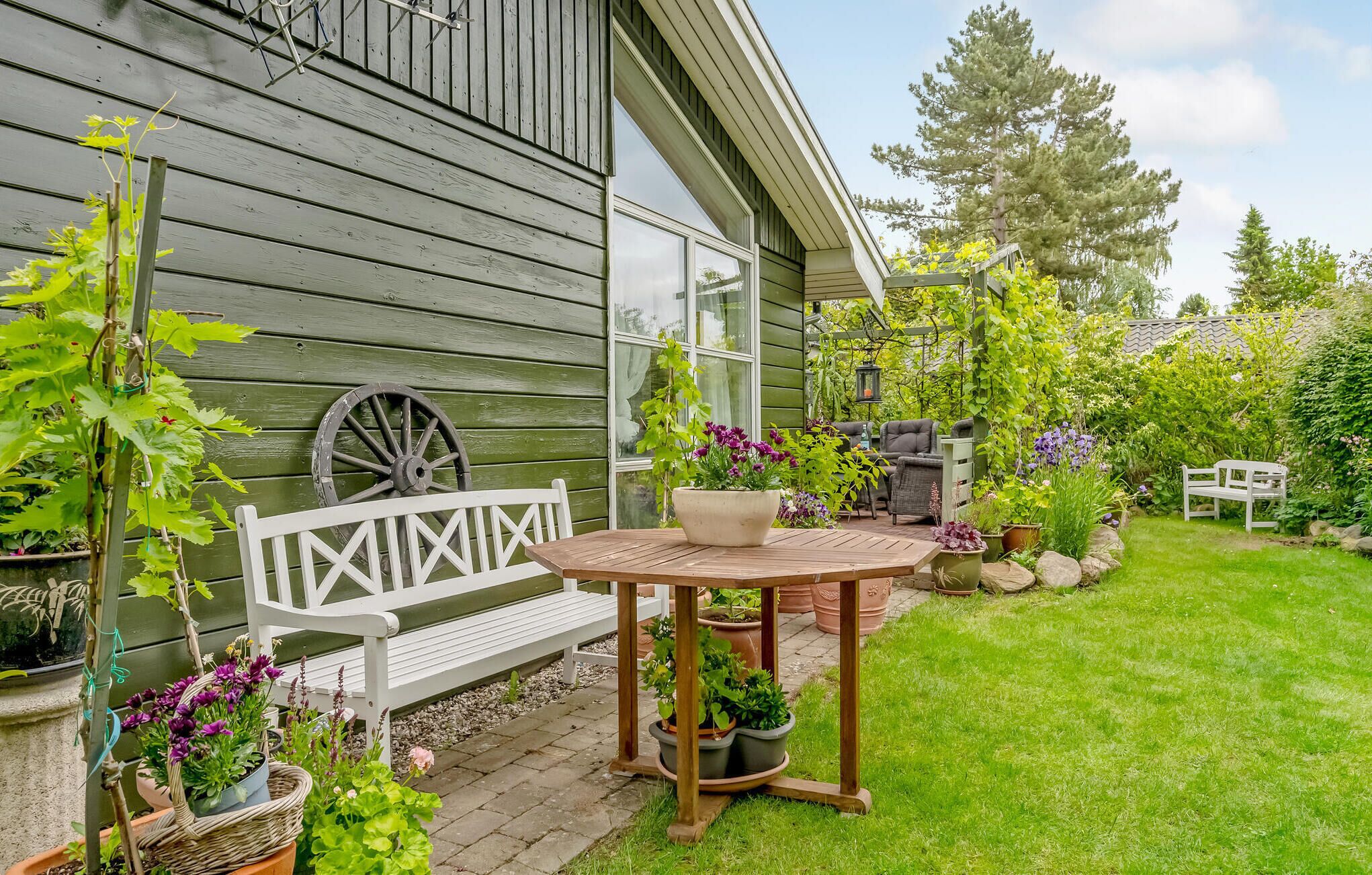 A wooden house with a porch, a white bench, a round table, and various potted plants.
