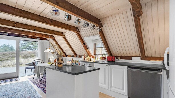 A modern kitchen with white cabinets, a black countertop, and a stainless steel dishwasher. There is a dining area with a table and chairs, and a view of the outdoors through a large window. - Lovely home in Rømø with kitchen (Rømø)