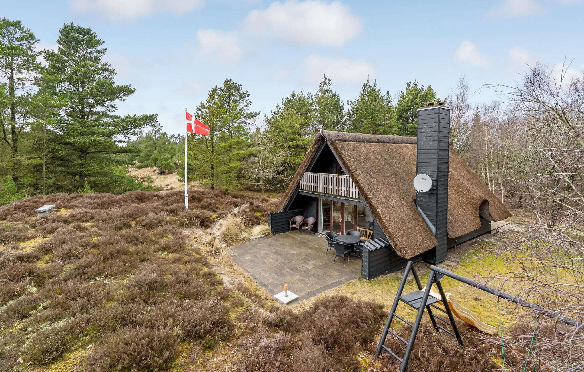 Een vakantiehuisje met rieten dak, een balkon en een vlaggenmast met vlag.