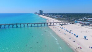A beach with a pier extending into the water, clear skies, and a few buildings in the background.