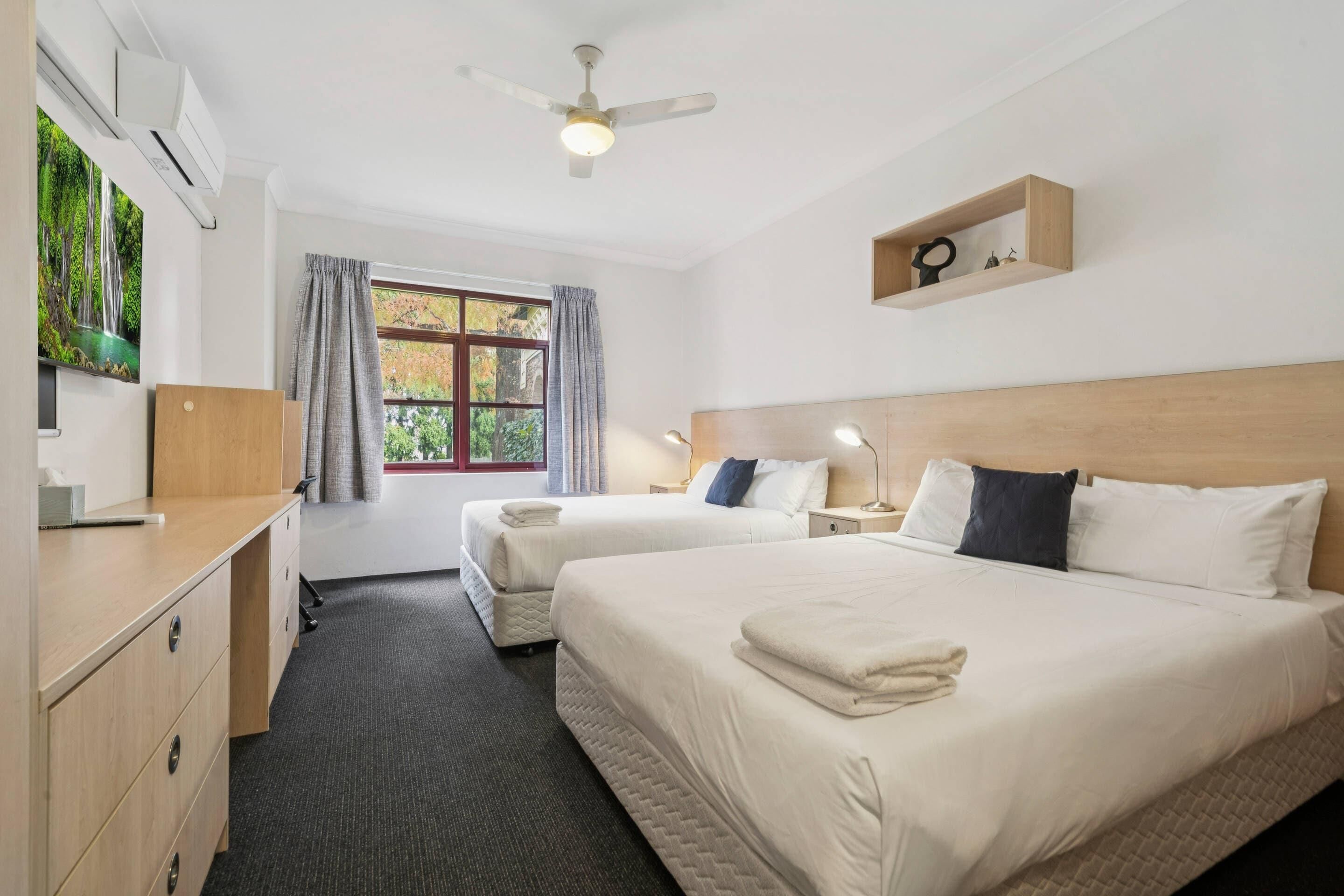 Une chambre d’hôtel avec deux lits, un bureau, un ventilateur de plafond, une fenêtre avec des rideaux et une étagère avec des objets décoratifs.