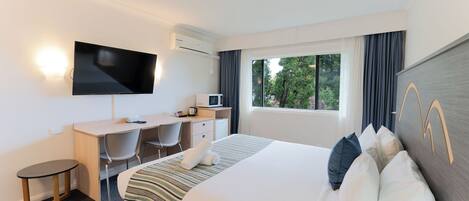 A hotel room with a bed, a desk, a TV, a window with curtains, and a view of trees.