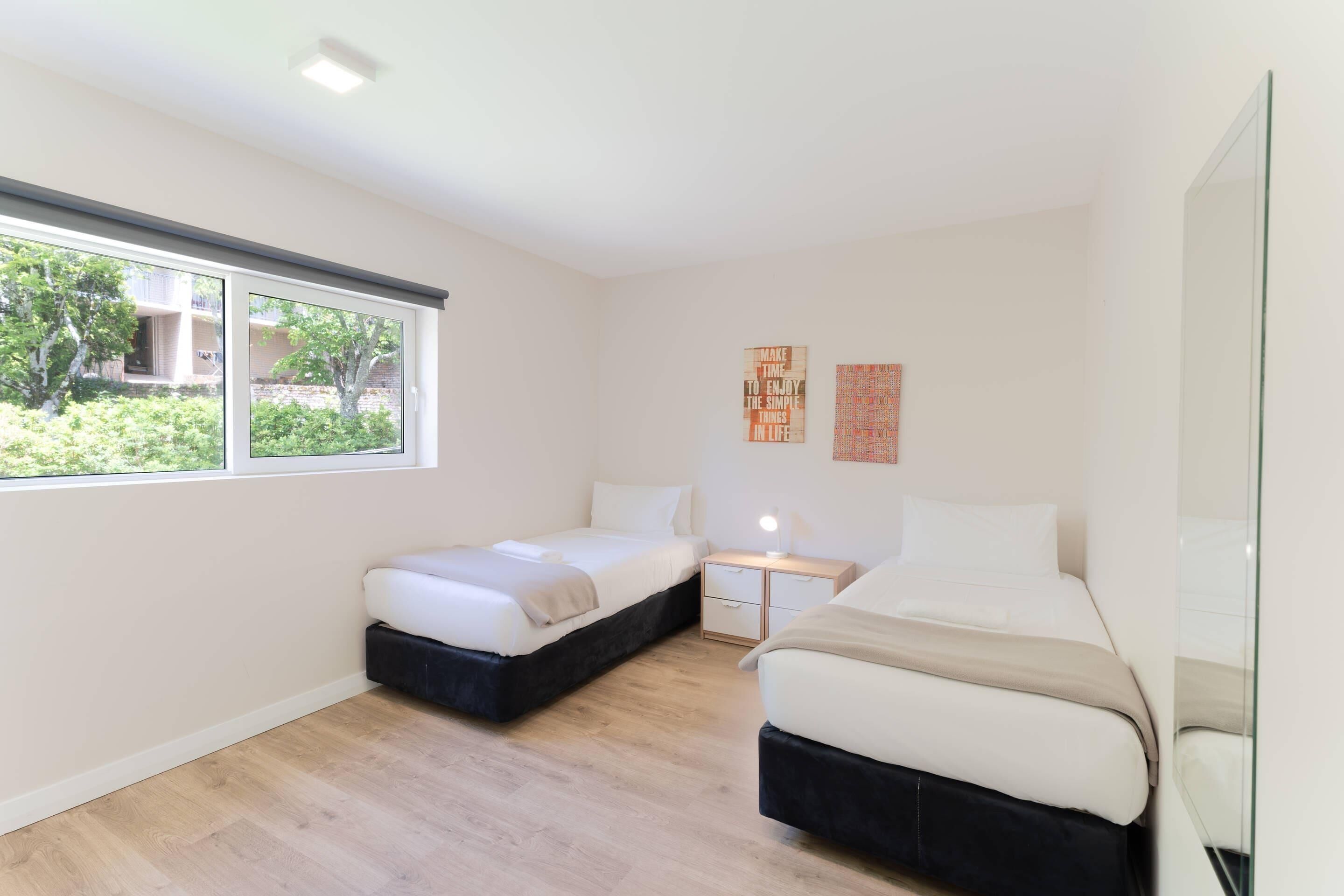 Une chambre avec deux lits, une fenêtre et une table de chevet avec une lampe.