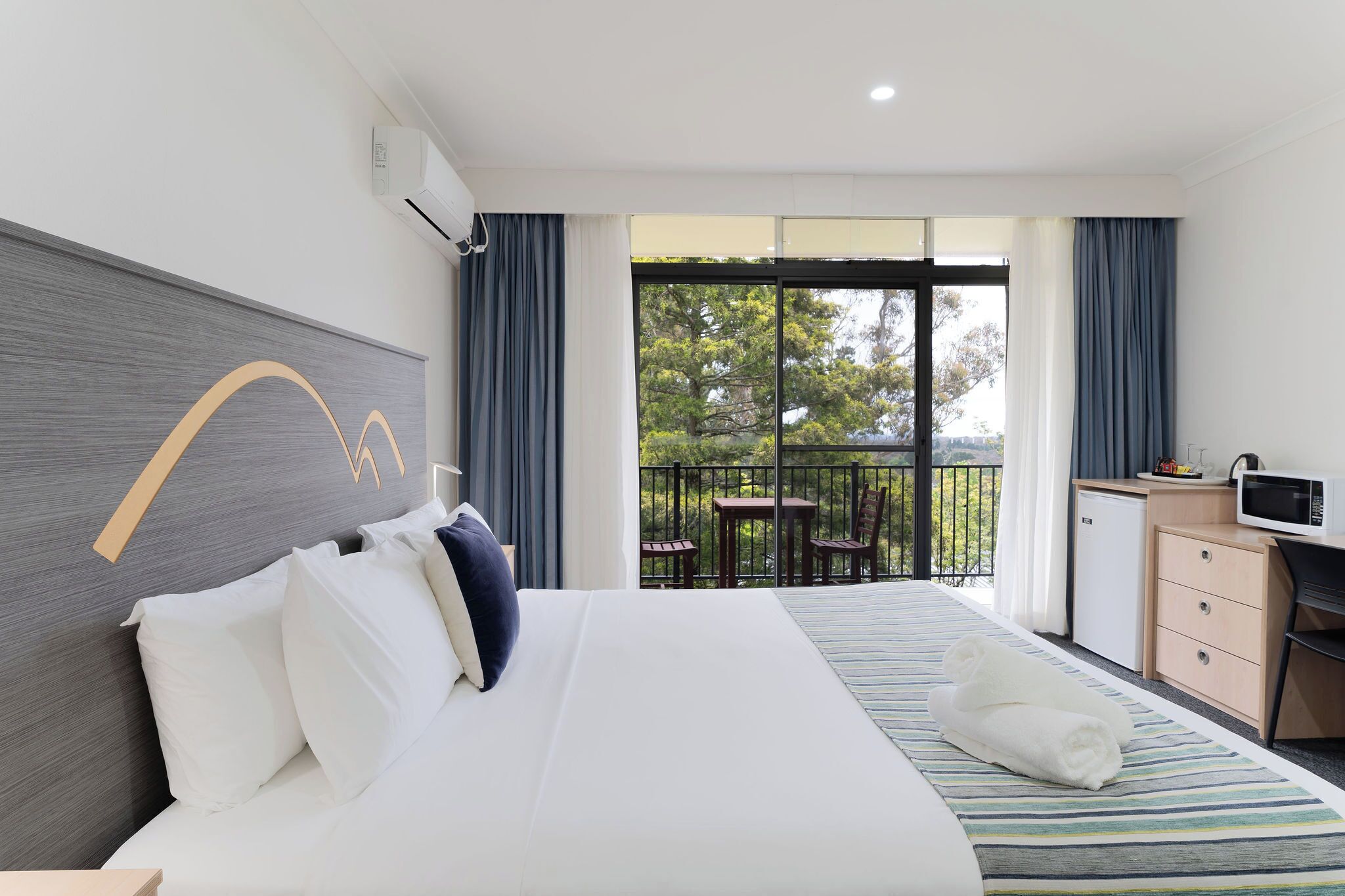 A hotel room with a large bed, a desk, a small fridge, and a balcony with a table and chairs.