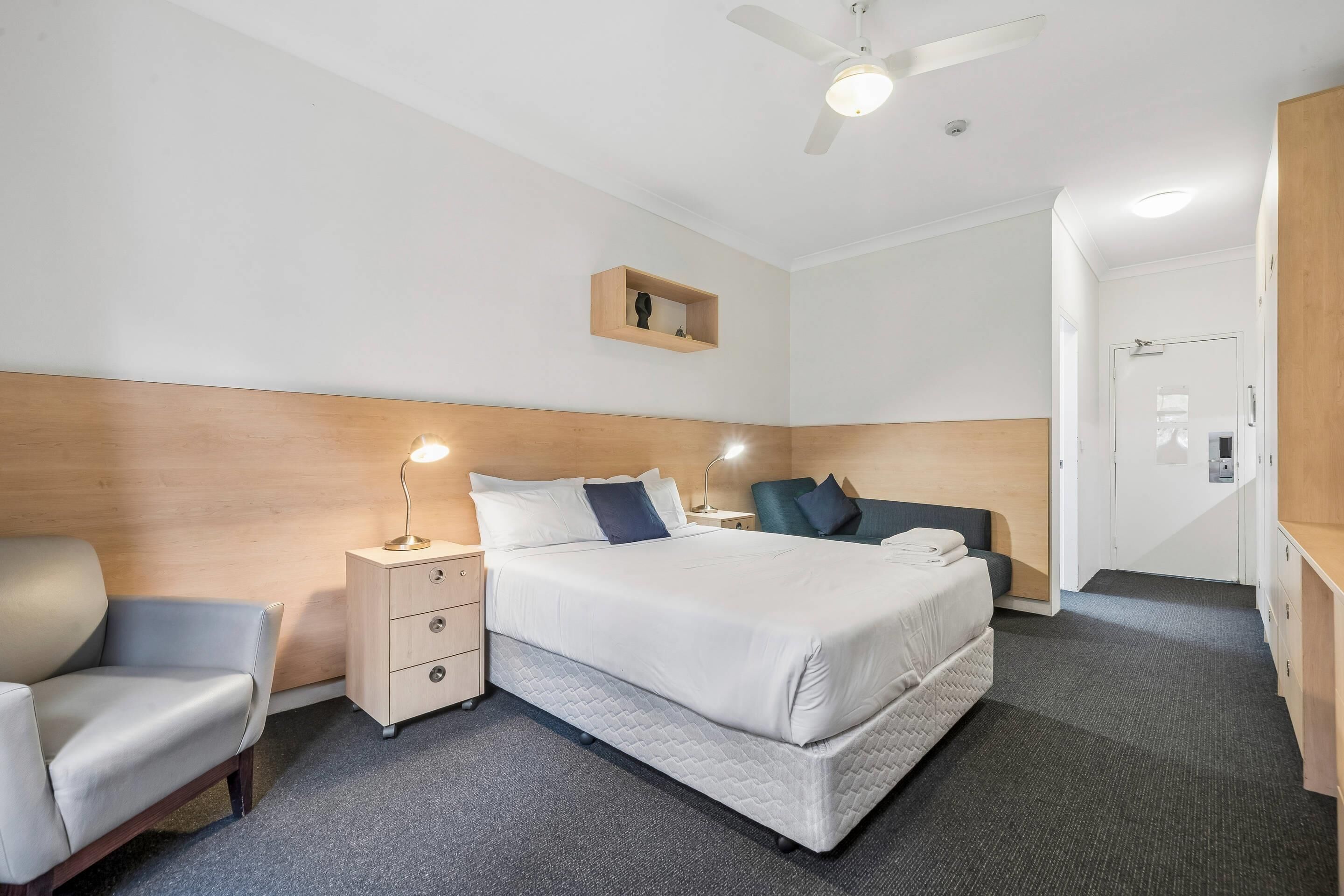 Habitación de hotel con cama, escritorio, silla, mesita de noche, ventilador de techo y estante de madera.