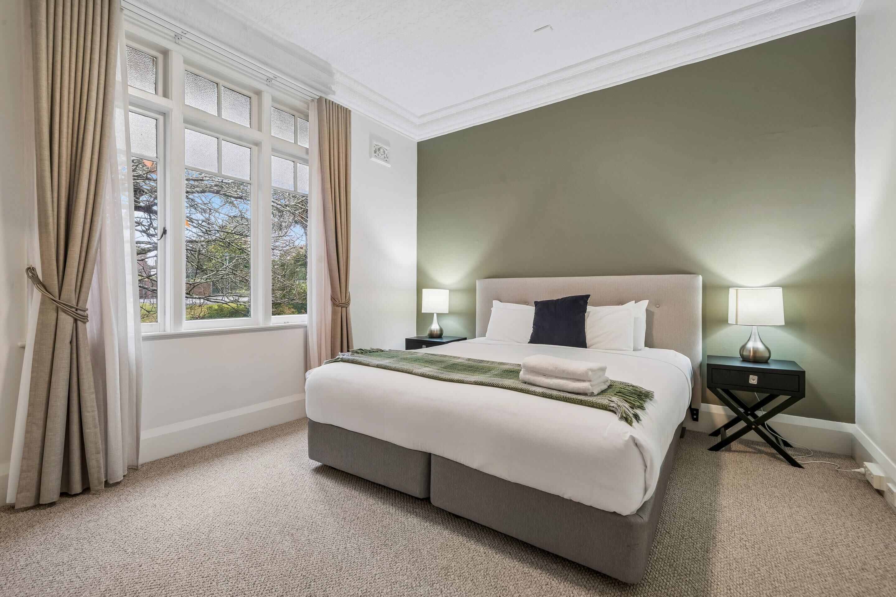 Een slaapkamer met een groot bed, twee nachtkastjes, een raam met gordijnen en uitzicht op bomen.
