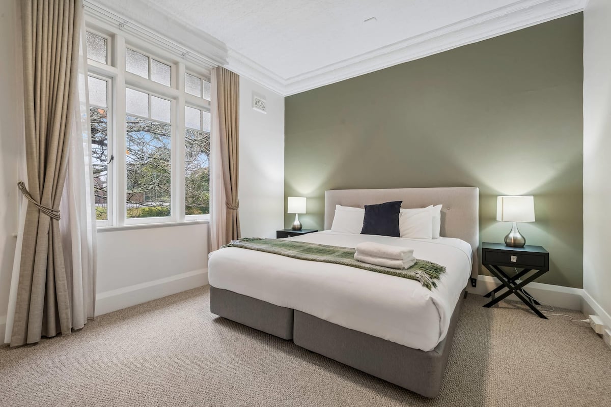 A bedroom with a large bed, two bedside tables, a window with curtains, and a view of trees.