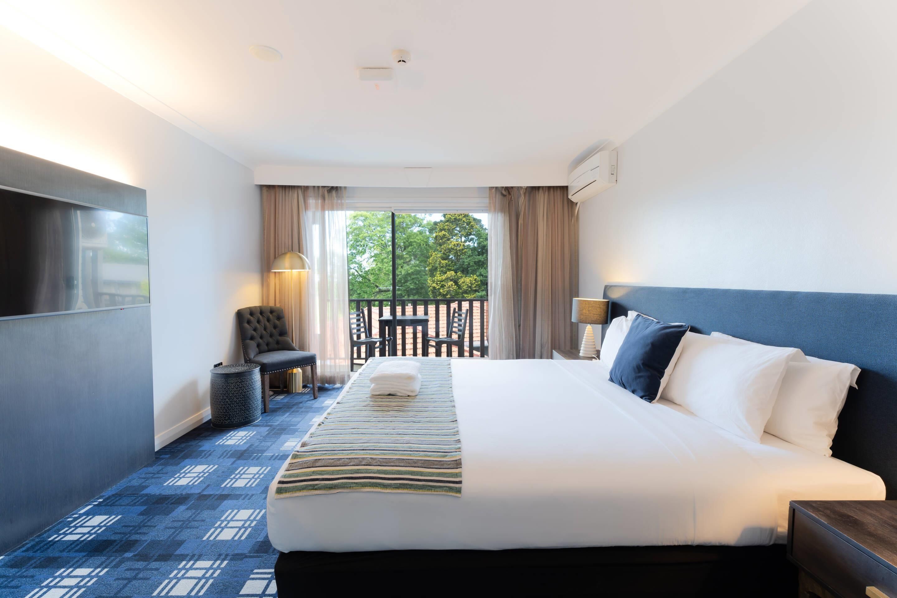 A hotel room with a large bed, a TV, a seating area with a chair and table, and a balcony with a view of trees.