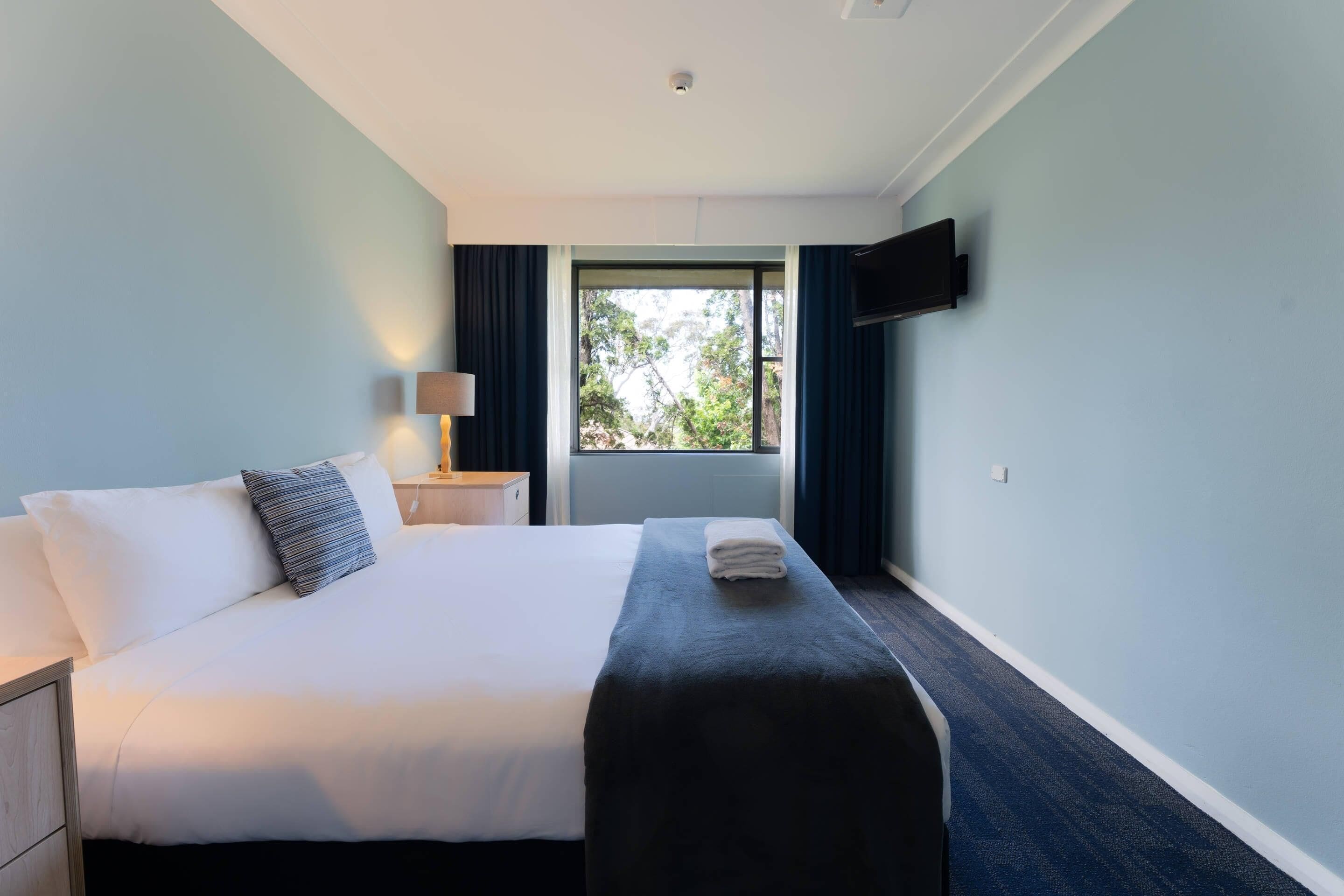 A hotel room with a bed, a bedside table, a lamp, a TV mounted on the wall, and a window with curtains.