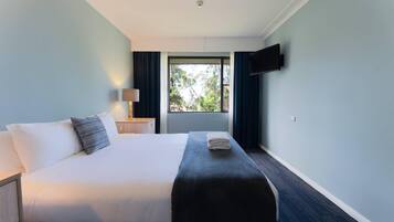 A hotel room with a bed, a bedside table, a lamp, a TV mounted on the wall, and a window with curtains.