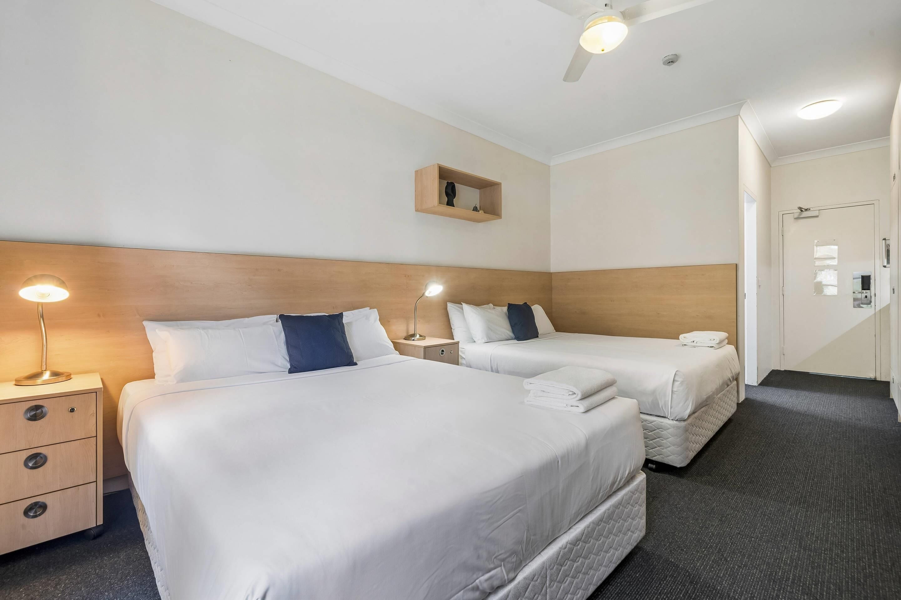 Habitación de hotel con dos camas, una mesita de noche con lámpara, un estante pequeño y una puerta.