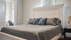 Une chambre à coucher avec un grand lit, une table de chevet, une chaise et une fenêtre avec des stores.