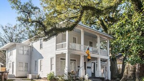 Une maison blanche à deux étages, dotée d’un balcon et entourée d’arbres.
