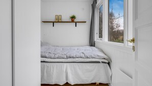 Ein kleines Schlafzimmer mit weißen Wänden, einem Bett, einem Fenster mit Vorhängen und einem Regal mit zwei gerahmten Bildern und einer Pflanze.