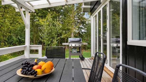 Ein überdachter Außenbereich mit Grill, Tisch mit Obst und Bank.