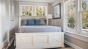 Une chambre à coucher avec un grand lit, une tête de lit blanche et deux fenêtres équipées de volets.