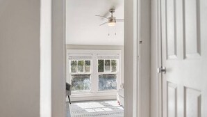 Un couloir mène à une pièce dotée d’un ventilateur de plafond, d’un tapis à motifs et d’une fenêtre avec des stores, offrant une vue sur l’extérieur.