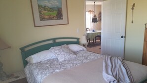 A neatly made bed with floral bedding and a blanket, a painting of a landscape on the wall, and a door leading to a dining area. - Wild Creek BnB, " Cottage " (Dyer)