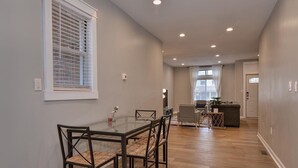 A spacious dining area with a glass table and chairs, a kitchen in the background, and a hallway leading to a door.