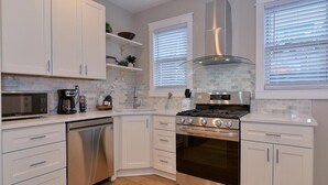 A modern kitchen with white cabinets, a stainless steel dishwasher, a gas stove, and a built-in oven. There are two windows with blinds, and a small shelf with decorative items.