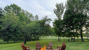 A circular stone fire pit with four Adirondack chairs around it, set in a grassy outdoor area with trees in the background. - Cozy, country cabin near Cooperstown (Richfield Springs)