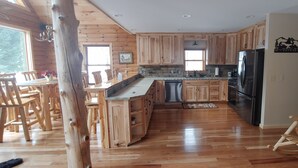 A spacious kitchen with wooden cabinets, a stainless steel refrigerator, and a dining area with wooden chairs. - Cozy, country cabin near Cooperstown (Richfield Springs)