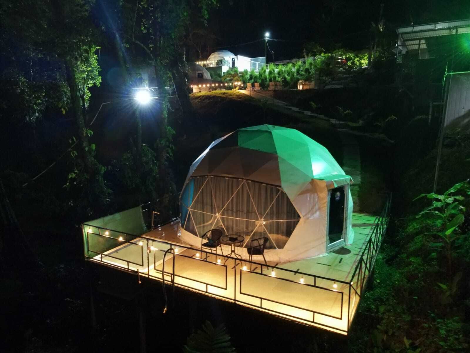 A geodesic dome structure with a glass floor, surrounded by trees and a lit pathway.