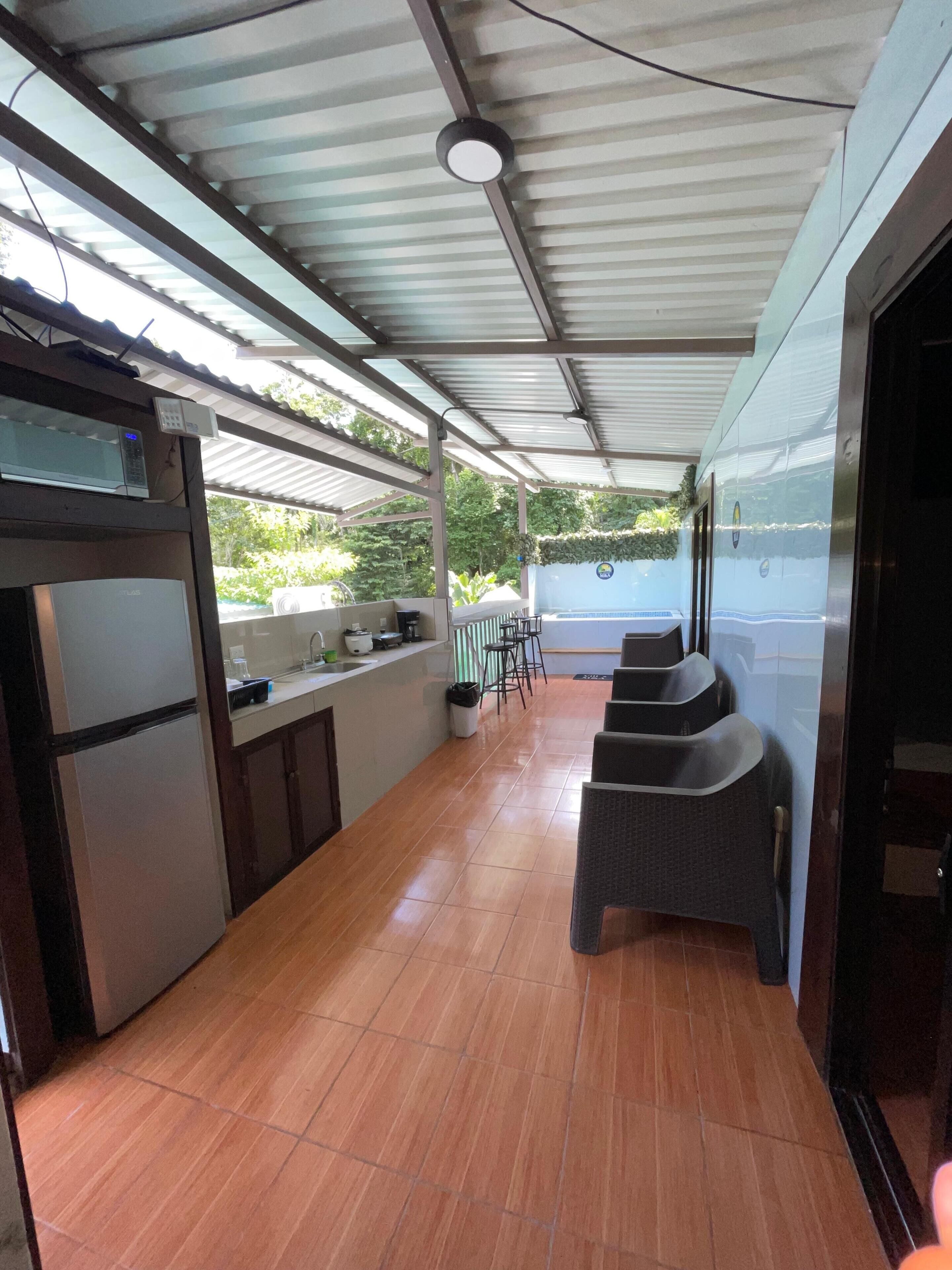 A covered outdoor area with a kitchen counter, a microwave, and a refrigerator. There are chairs and a table in the background.