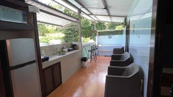 A covered outdoor area with a kitchen counter, a microwave, and a refrigerator. There are chairs and a table in the background.