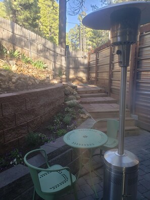 A patio with a metal heater, two green chairs, a small table, a brick wall, a wooden fence, and a stone retaining wall. - The Tiny  Basecamp  (Flagstaff)