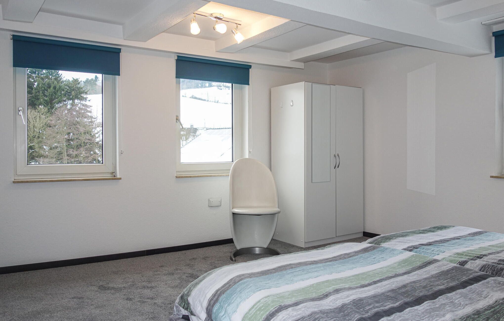 Une chambre à coucher comprenant un lit, une armoire, une chaise et deux fenêtres équipées de stores.